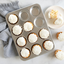 Nordic Ware aluminum 12-cavity muffin pan with vanilla frosted cupcakes, one cupcake on white plate