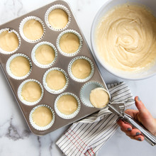 Person pouring vanilla muffin batter into paper liners in Nordic Ware aluminum 12-cup muffin pan