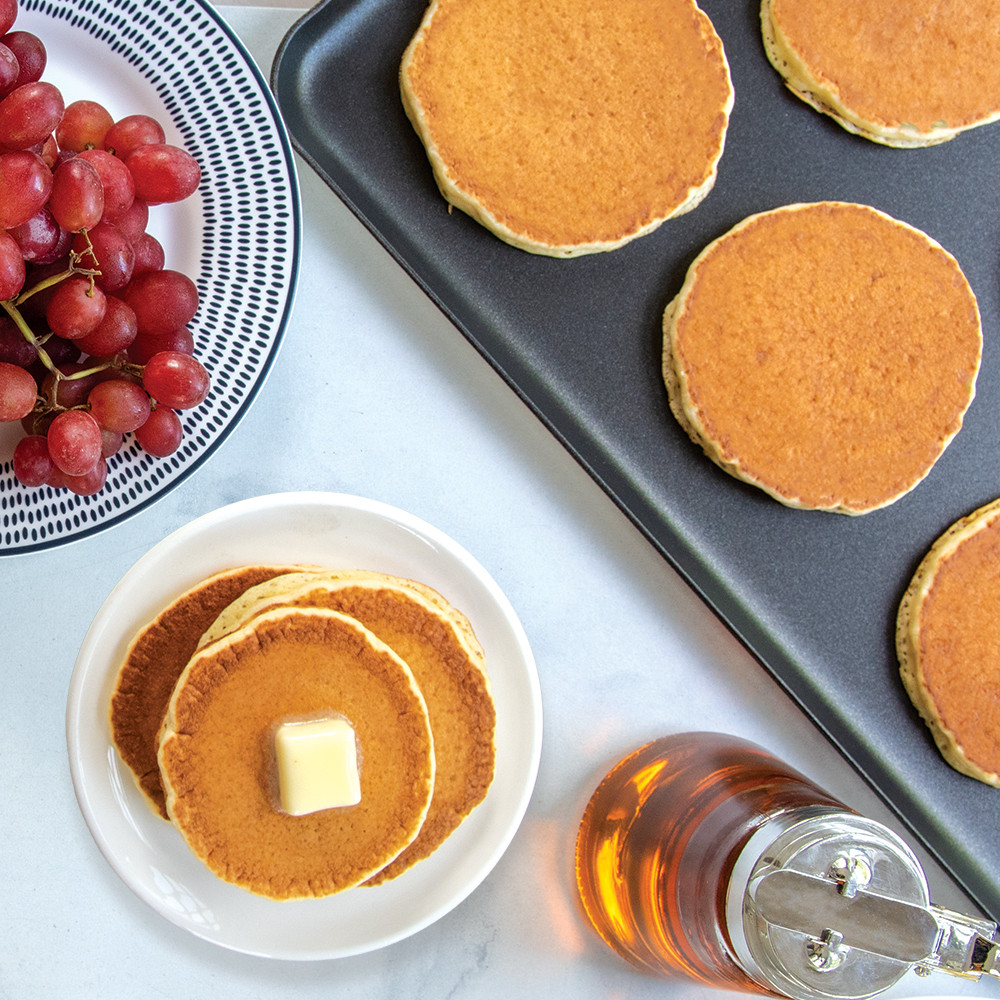 Golden pancakes cooking on Nordic Ware two-burner griddle with stack of finished pancakes, berries and syrup nearby