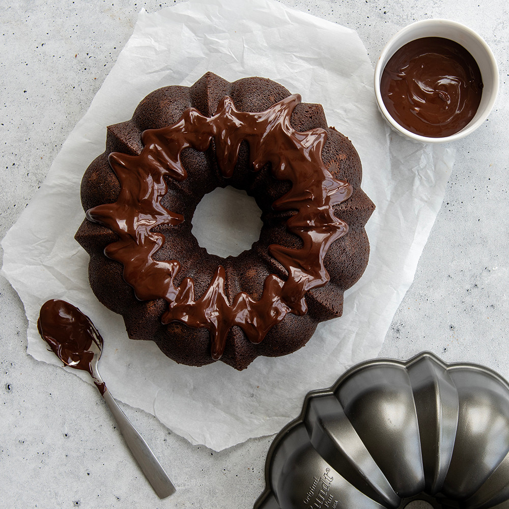 Chocolate bundt cake with glossy ganache glaze beside Nordic Ware aluminum ProCast bundt pan with fluted design
