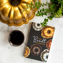 Nordic Ware Anniversary Bundt pan with The Bundt Cookbook, coffee cup and plant on white marble surface