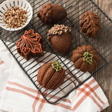 Six golden pumpkin-shaped cakes on wire cooling rack, decorated with orange, yellow, and green frosting, one topped with chopped nuts