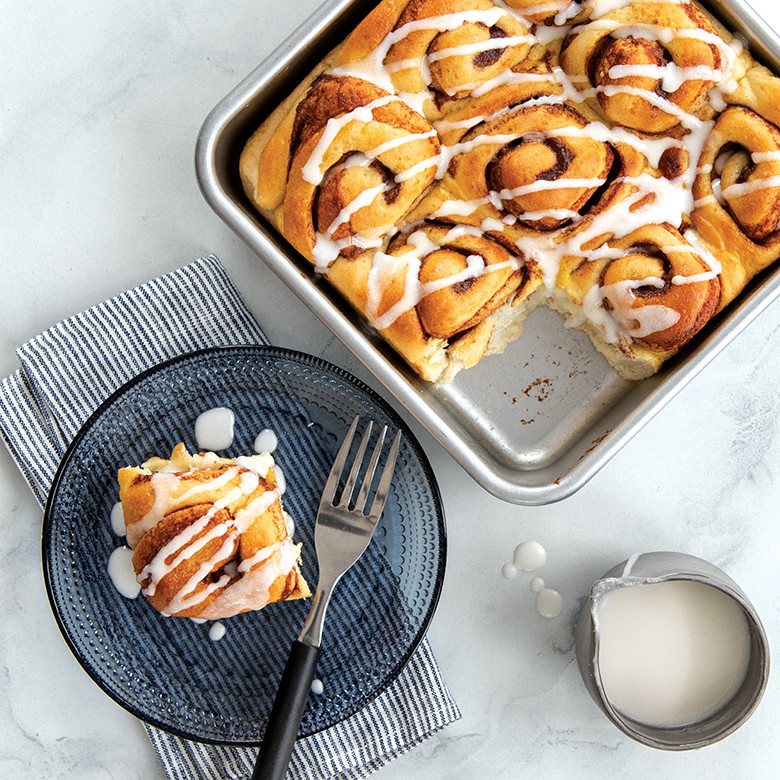 Nine golden cinnamon rolls baked in Nordic Ware aluminum square cake pan, one roll removed and plated nearby