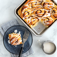 Nine golden cinnamon rolls baked in Nordic Ware aluminum square cake pan, one roll removed and plated nearby