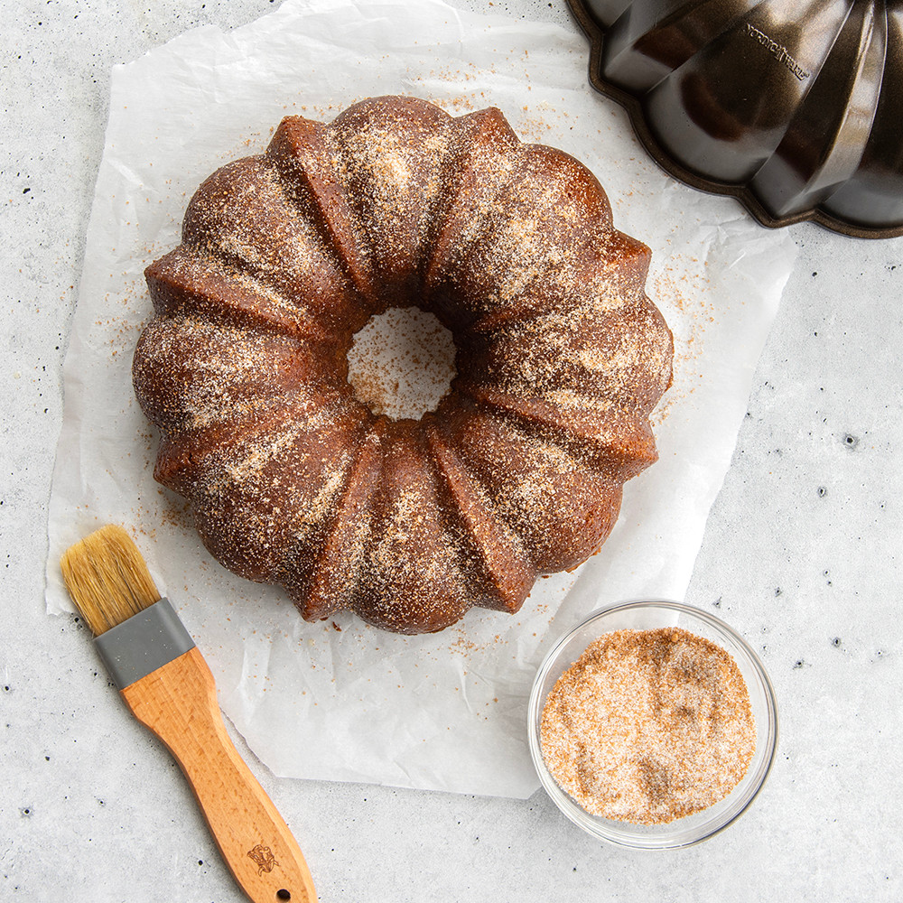 Golden bundt cake with cinnamon sugar coating on parchment beside bronze-finished Nordic Ware bundt pan
