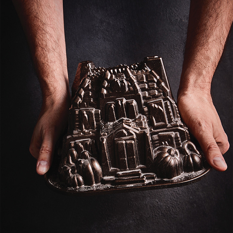 Two hands holding Nordic Ware Haunted Manor bundt pan, cast aluminum with intricate gothic architectural details
