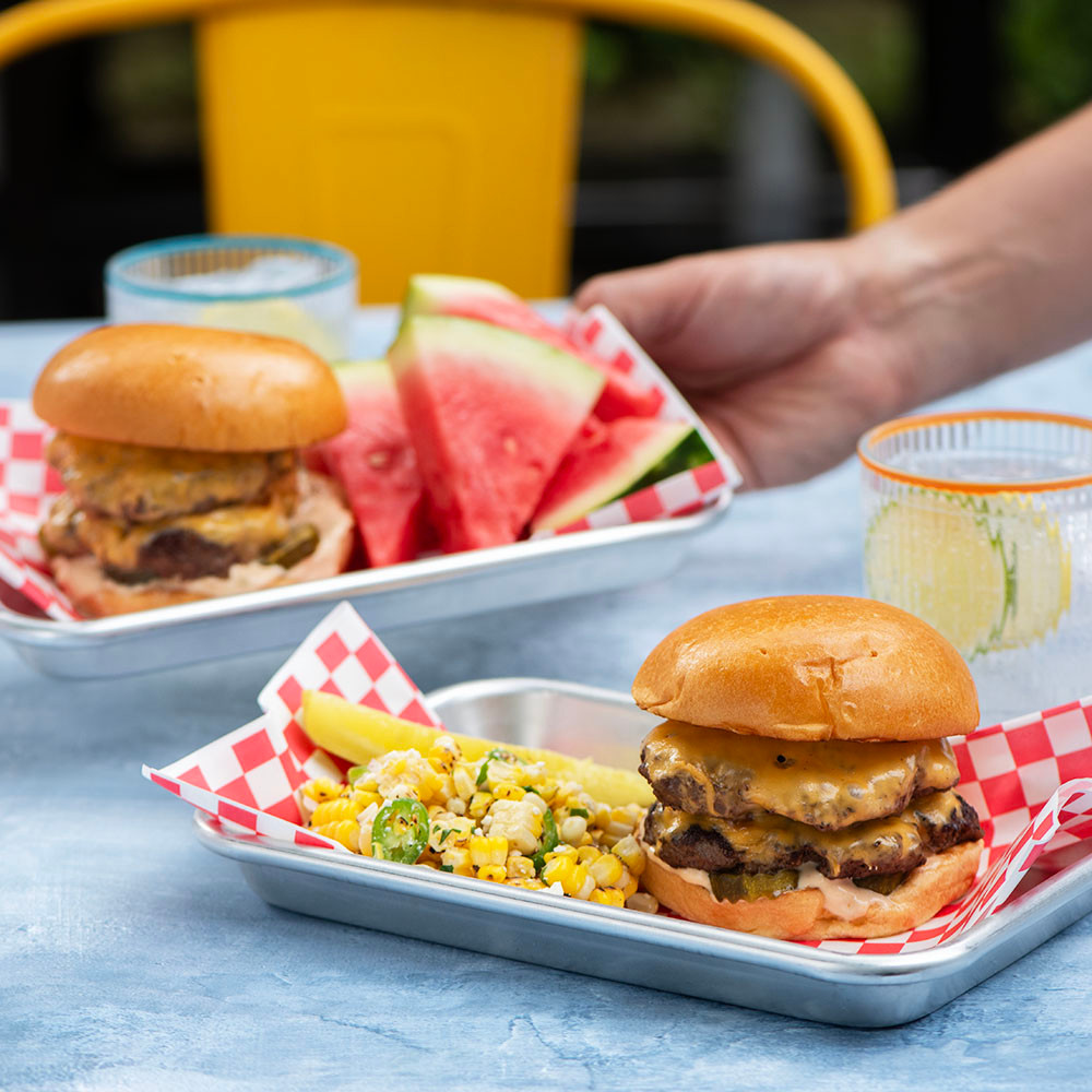 Two Nordic Ware aluminum burger serving trays with burgers, corn salad, and watermelon, hand reaching for one tray
