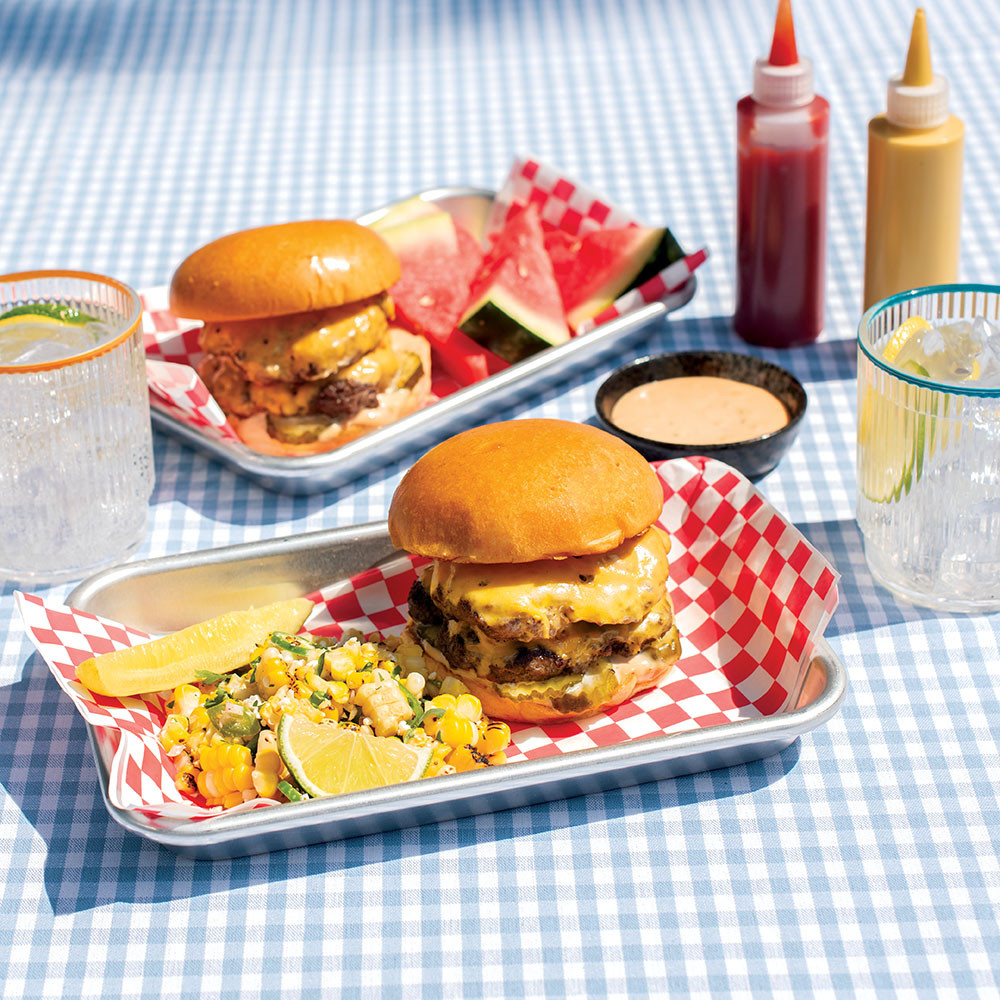 Two Nordic Ware aluminum burger serving trays with cheeseburgers, corn salad, and watermelon on checkered tablecloth