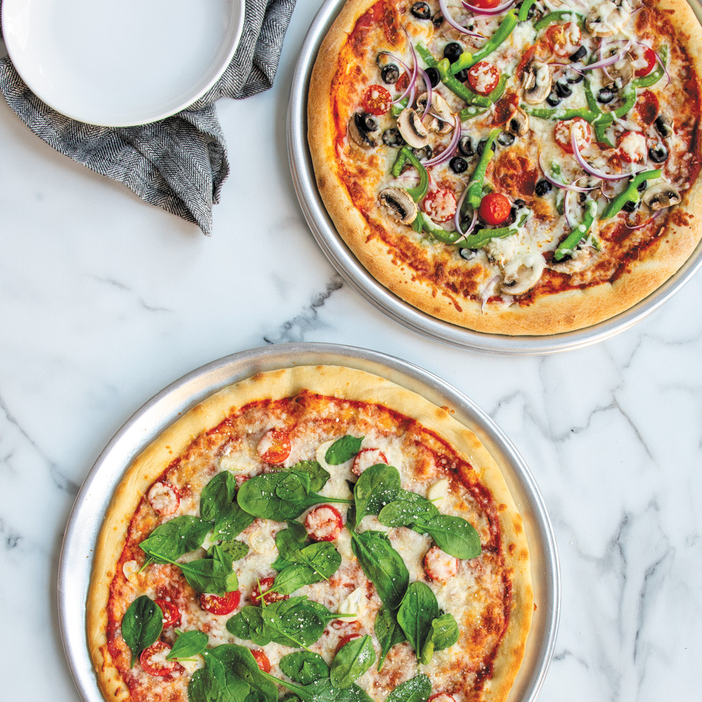 Two Nordic Ware aluminum pizza pans with baked pizzas - one traditional smooth, one perforated crisper pan