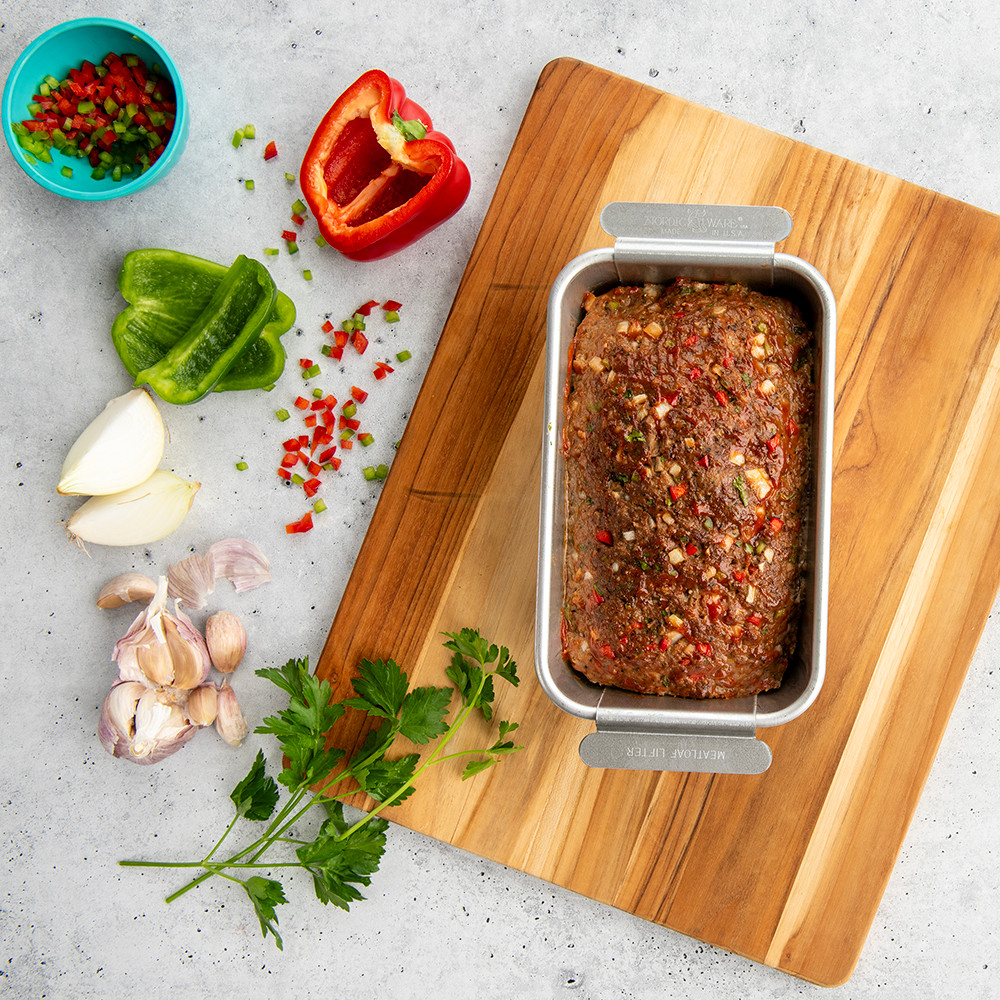 Nordic Ware aluminum meatloaf pan with baked seasoned meatloaf, surrounded by fresh vegetables and herbs