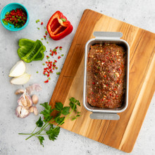 Nordic Ware aluminum meatloaf pan with baked seasoned meatloaf, surrounded by fresh vegetables and herbs