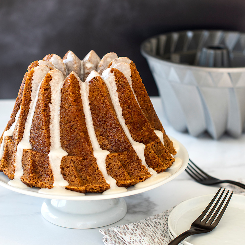Golden carrot bundt cake with white glaze drizzled down fluted sides, Nordic Ware Kugelhopf pan behind