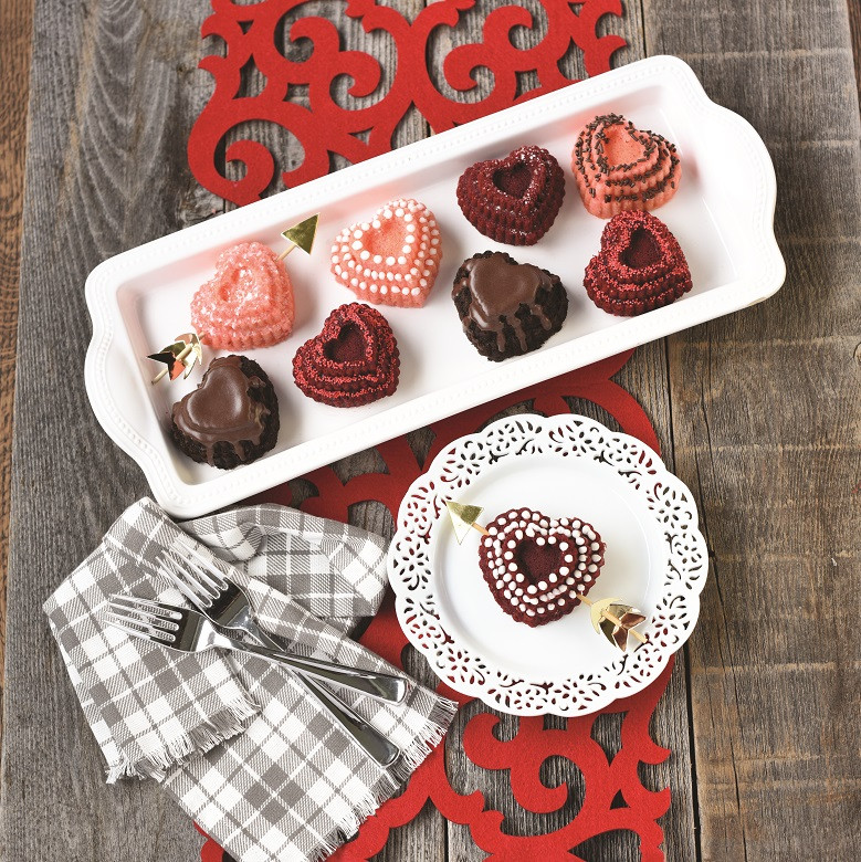 Heart-shaped cakelets with chocolate glaze and decorative frosting on white serving tray, one plated with gold arrow