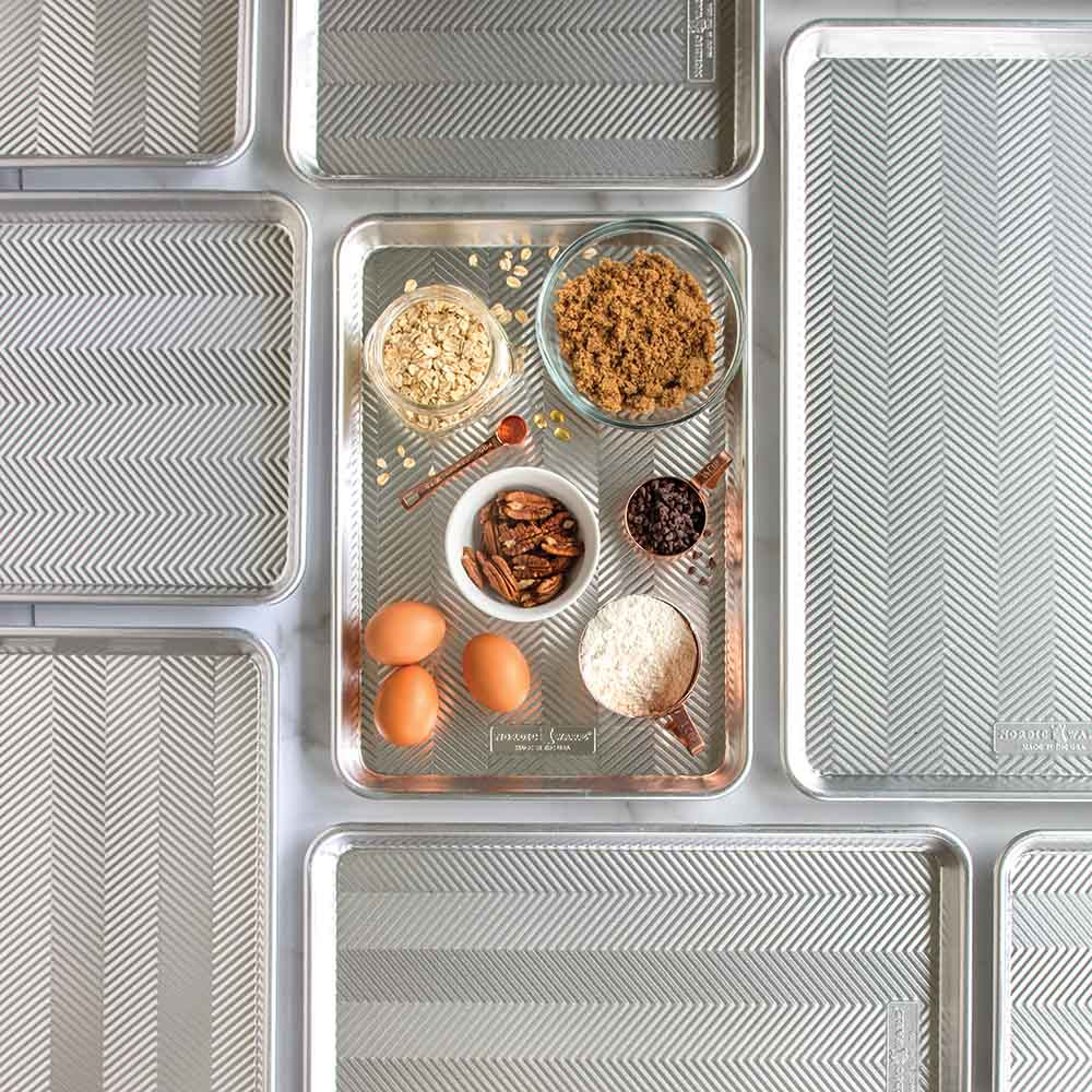 Nordic Ware Prism aluminum baking sheets with textured surface, featuring baking ingredients including eggs, flour, nuts, and chocolate chips