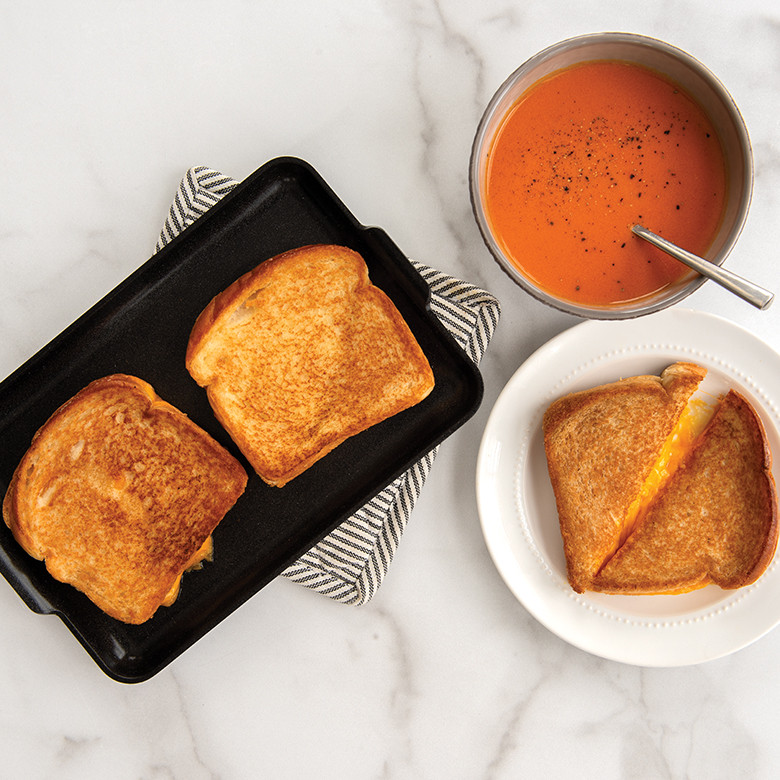 Nordic Ware black non-stick mini griddle with two golden grilled cheese sandwiches, served with tomato soup