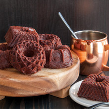 Six chocolate bundt cakes with geometric ridged patterns on wooden serving board, copper pot with spoon nearby