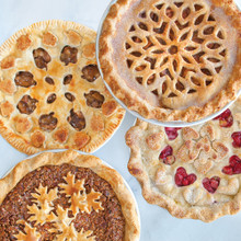Four golden-brown baked pies in Nordic Ware aluminum pans featuring decorative lattice, leaf, and heart crust designs