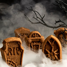 Golden baked tombstone-shaped cakes with detailed engravings, surrounded by spooky smoke and bare branches