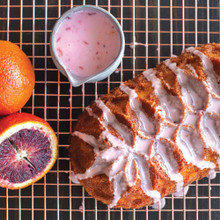 Sliced blood orange loaf cake with pink glaze drizzle on cooling rack, bowl of glaze and fresh oranges alongside
