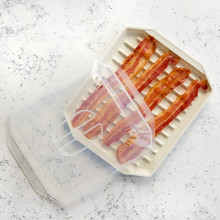 Nordic Ware compact bacon tray with clear lid, showing crispy and raw bacon strips on ridged cream-colored base