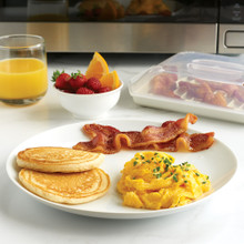 Nordic Ware white compact bacon tray with clear lid containing cooked bacon strips beside breakfast plate