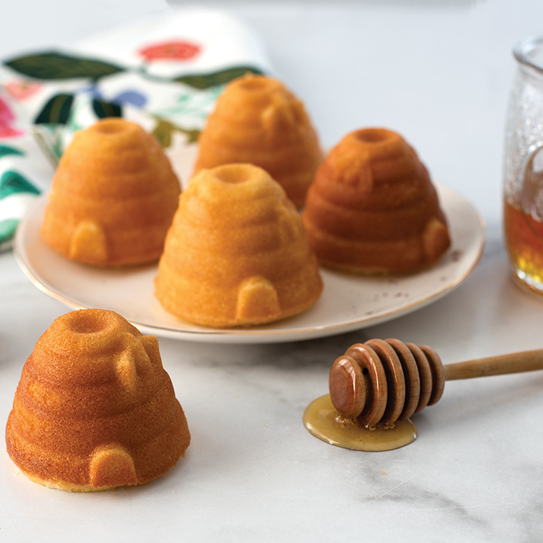 Golden-brown beehive cakelets with detailed ridged texture, four on white plate and one on marble surface with honey dipper