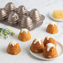 Golden-brown beehive cakelets with detailed ridged texture, four on white plate and one on marble surface with honey comb and flowers