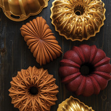 Multiple golden bundt cakes in Nordic Ware decorative aluminum pans showing intricate fluted and geometric designs