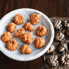 Golden mini bundt cakes on white plate next to Nordic Ware aluminum Autumn Delights cakelet pan with leaf and acorn molds