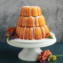 Three-tiered golden bundt cakes with white glaze drizzle on white pedestal stand, decorated with grapes