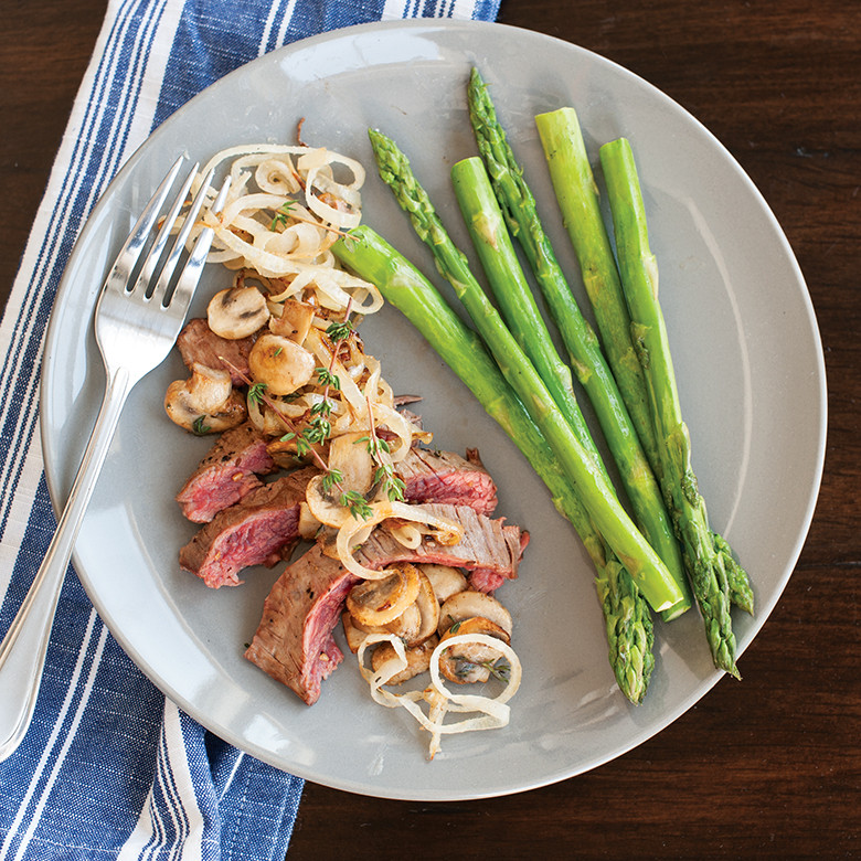 Sliced medium-rare steak with sautéed mushrooms and onions, served with fresh asparagus spears on white plate