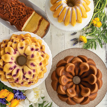 Four baked bundt cakes with floral designs: chocolate lotus cake, glazed yellow cake, petal-shaped cake with purple flowers, and bronze heritage bundt
