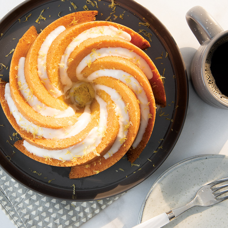 Golden bundt cake with white glaze and orange zest on dark plate, showing Nordic Ware Heritage pan's fluted design