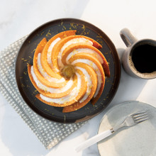 Golden bundt cake with white glaze on dark plate, Nordic Ware Heritage aluminum bundt pan and coffee cup nearby