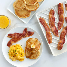 Nordic Ware aluminum slanted bacon tray with crispy bacon strips, plus plates of pancakes and eggs with orange juice