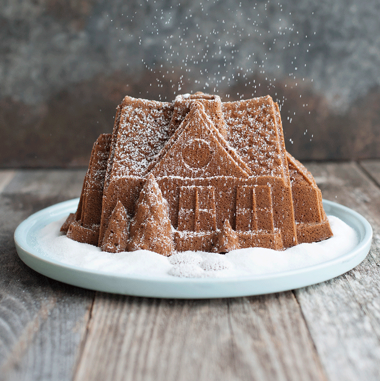Powdered sugar falling onto golden gingerbread bundt cake shaped like detailed house with windows and door