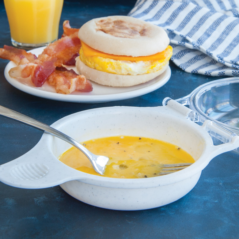 Raw egg cooking in Nordic Ware Egg 'N Muffin aluminum breakfast pan with assembled sandwich and bacon on plate
