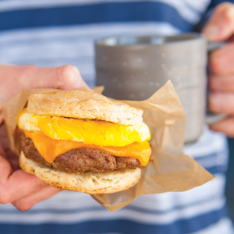 Hand holding breakfast sandwich with melted cheese and sausage patty on split biscuit in parchment wrapper