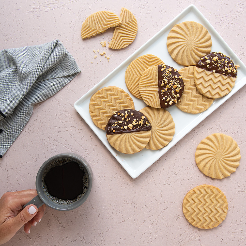 Geometric-stamped cookies, some chocolate-dipped, arranged on white plate with hand holding coffee cup