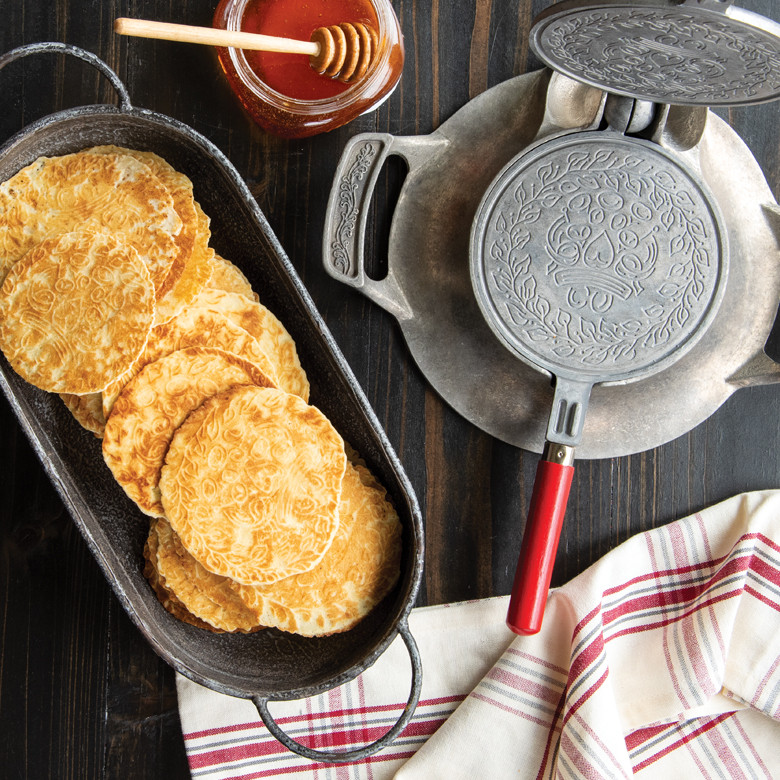 Nordic Ware aluminum krumkake iron open beside golden pizzelles arranged in rows, honey bowl in background