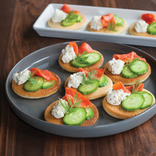 Small golden pancakes on white platter topped with smoked salmon, cucumber slices, dill and cream cheese