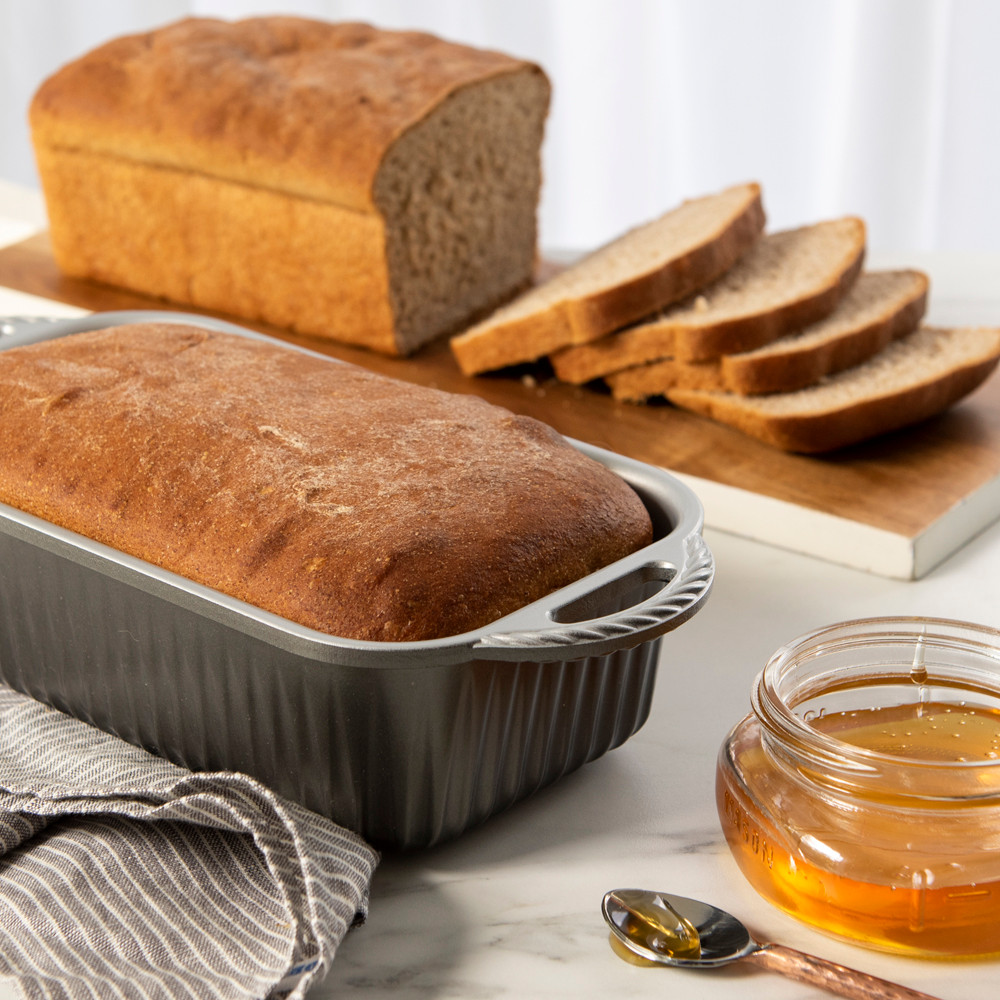 Golden-brown baked loaf in Nordic Ware aluminum Classic Loaf Pan with sliced loaf on cutting board nearby