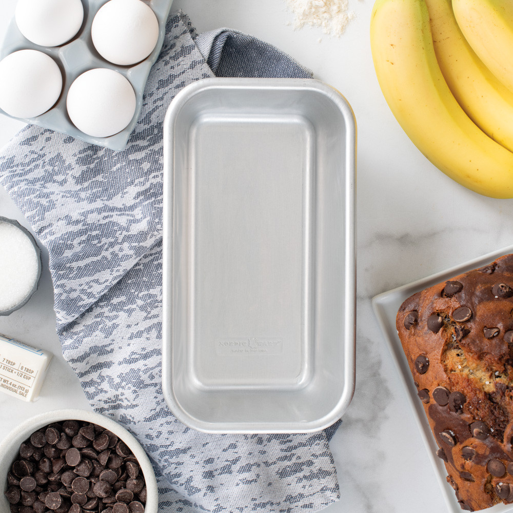 Nordic Ware natural aluminum 1-pound loaf pan with baking ingredients including flour, eggs, and butter arranged around it