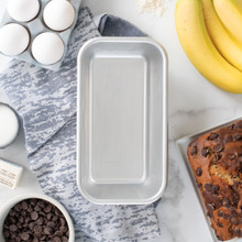 Nordic Ware natural aluminum 1-pound loaf pan with baking ingredients including flour, eggs, and butter arranged around it