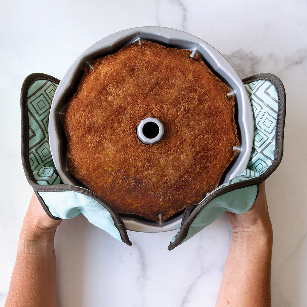 Two colorful Nordic Ware bundt-shaped hot pads holding aluminum bundt pan with golden baked cake