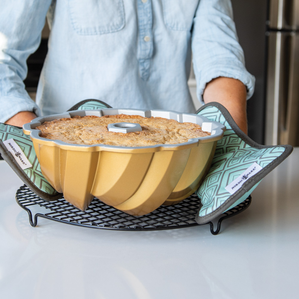 Woman using Nordic Ware bundt-shaped hot pads to transfer golden bundt cake in aluminum pan to cooling rack
