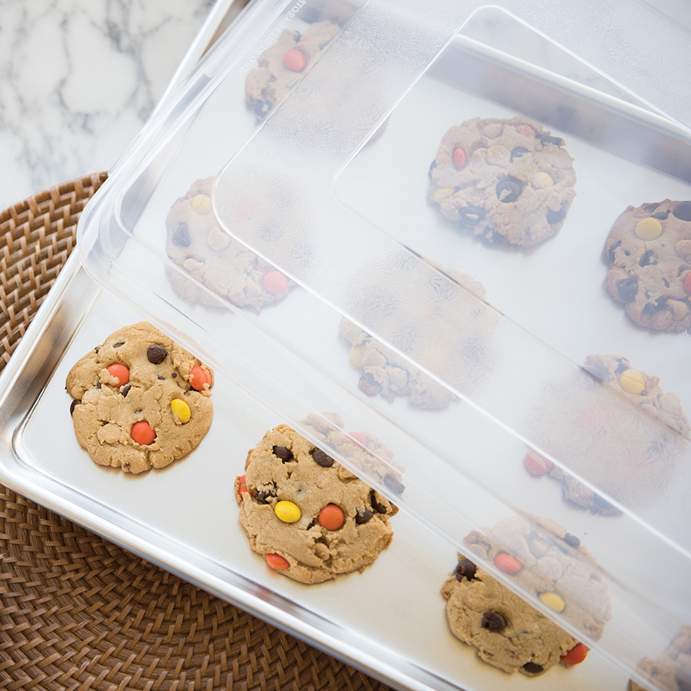 Nordic Ware aluminum half sheet pan with clear lid, containing golden baked chocolate chip cookies