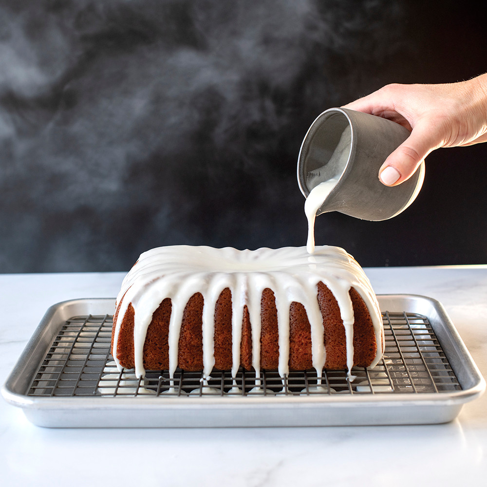 Hand drizzling white glaze over golden loaf cake on wire cooling grid, showing quarter sheet size