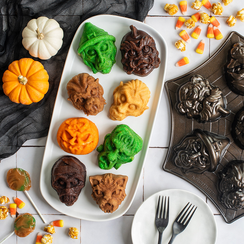 Multi color Monster Mask Cakelets on plate next to pan on tile backdrop, assorted Halloween candy spread throughout