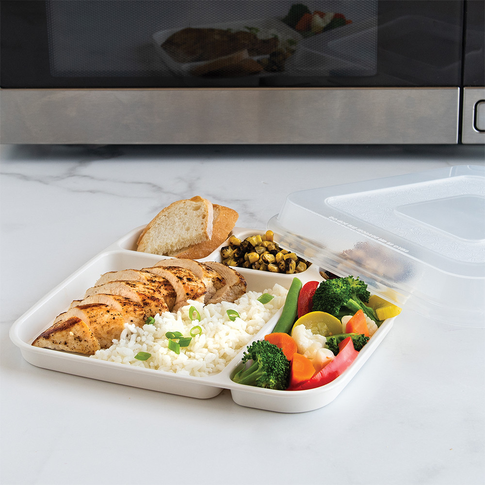 Nordic Ware aluminum divided dinner tray with clear lid partially open, containing leftover food portions, positioned near microwave